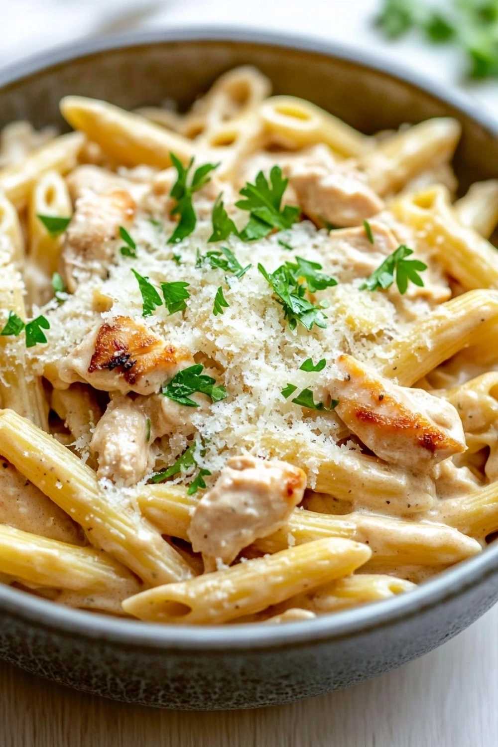 One pot creamy garlic chicken pasta - the image is a close-up of a bowl of penne pasta. the pasta is cooked al dente and is topped with chunks of chicken and grated parmesan cheese. the dish is garnished with fresh parsley. the bowl is made of ceramic and is sitting on a wooden table. the background is blurred, but it appears to be a kitchen countertop.