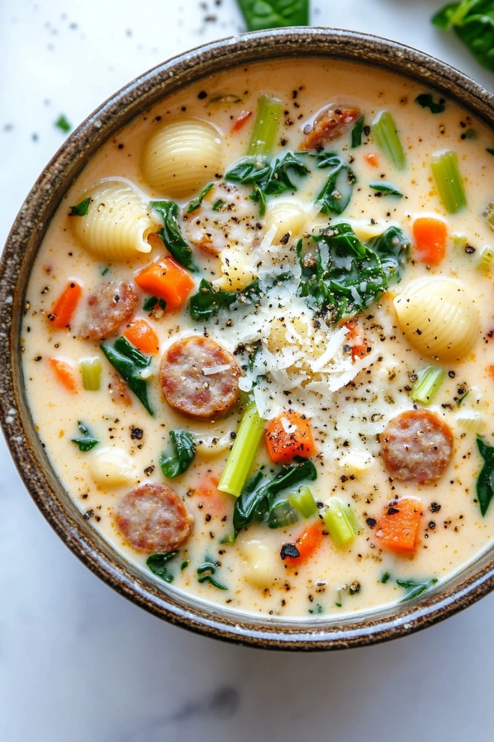 One pot creamy italian sausage soup - the image is a close-up of a bowl of soup. the soup appears to be a creamy yellow color with chunks of sausage, carrots, celery, and other vegetables mixed in. the bowl is made of ceramic and has a brown rim. there are also some crumbled parmesan cheese scattered throughout the soup, adding a crunchy texture. the background is a white marble countertop with a few sprigs of fresh basil scattered around.