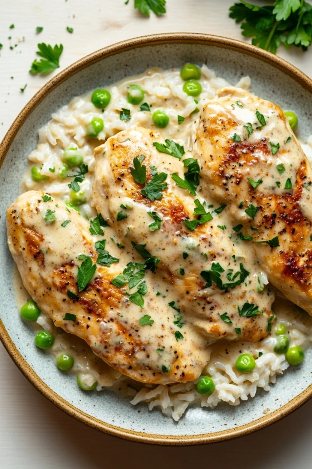 One pot creamy parmesan chicken and rice - the image shows a plate of food with two pieces of chicken breasts covered in a creamy sauce. the chicken breasts are golden brown and appear to be seasoned with herbs and spices. the sauce is a light yellow color and is drizzled over the top of the chicken breasts. the dish is served on a bed of white rice with small green peas scattered throughout. the plate is garnished with a sprig of fresh parsley. the background is a white tablecloth with a few sprigs of parsley scattered around.
