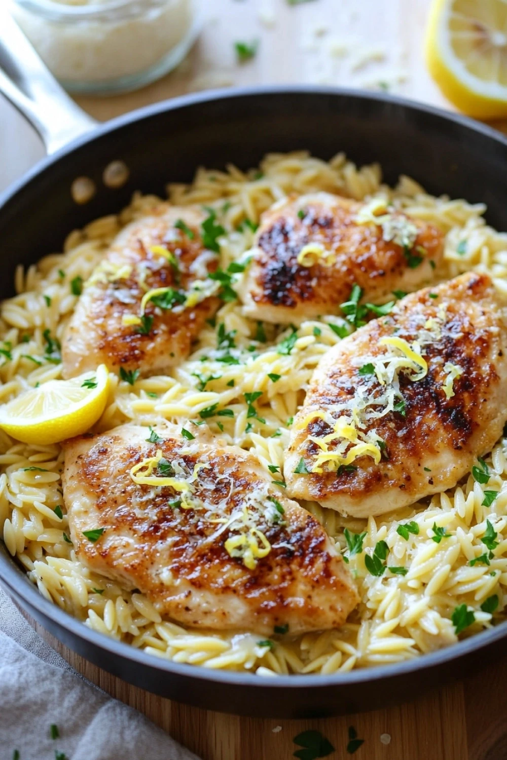 One skillet lemon butter chicken and orzo - the image shows a black cast iron skillet with four pieces of grilled chicken on top of a bed of yellow pasta. the chicken appears to be seasoned with herbs and spices, and there are two lemon wedges on either side of the skillet. the skillet is sitting on a wooden cutting board with a white cloth napkin underneath. in the background, there is a small jar of creamy sauce and a few sprigs of parsley. the overall color scheme of the image is warm and inviting.