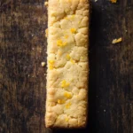 Orange shortbread cookie sticks gluten free - the image is a close-up of a rectangular piece of bread on a dark wooden surface. the bread appears to be freshly baked and has a golden-brown color. the edges of the bread are slightly crumbly and there are small pieces of orange zest scattered around the edges. the background is blurred, making the bread the focal point of the image.
