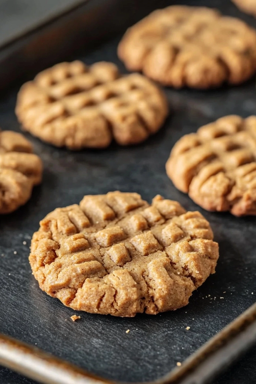Peanut butter cookie for dogs - the image shows a baking tray with freshly baked cookies on it. the cookies are round and golden brown in color, with a criss-cross pattern on top. they are arranged in a single layer on the tray, with some overlapping each other. the tray is lined with parchment paper, and there are a few crumbs scattered around the cookies. the background is dark, making the cookies stand out.
