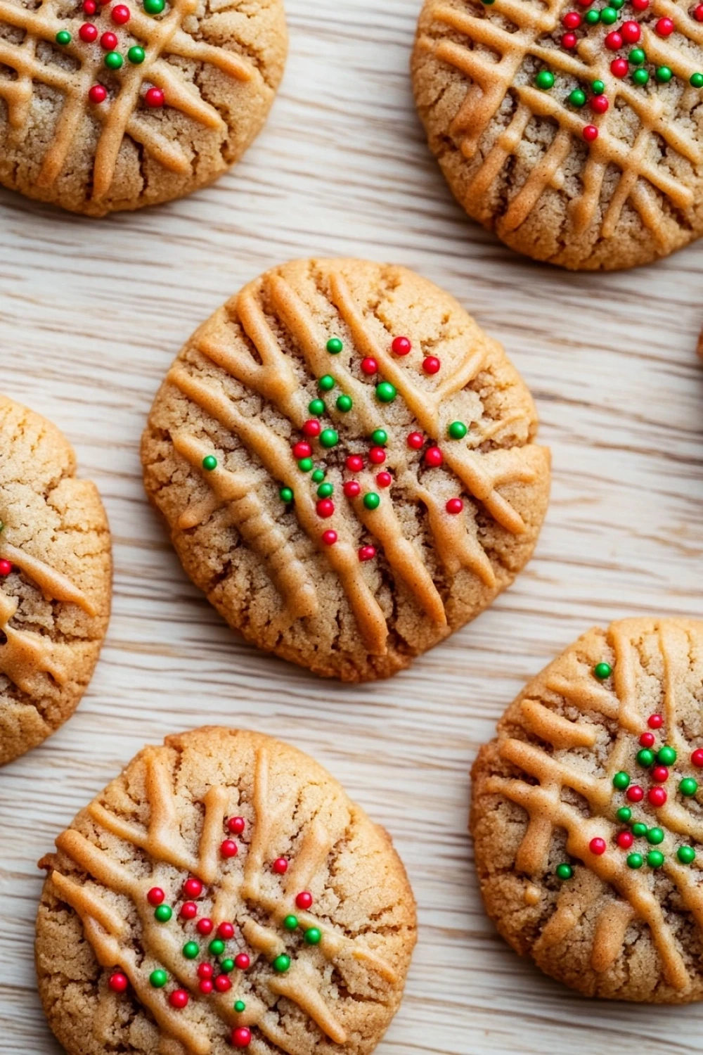 Peanut butter xmas cookie - the image shows a group of freshly baked cookies on a white wooden surface. the cookies are round and golden brown in color, with a criss-cross pattern on top. each cookie is decorated with colorful sprinkles in shades of red, green, and yellow. the sprinkles are arranged in a symmetrical pattern, with some overlapping each other. the background is blurred, but it appears to be a kitchen countertop.