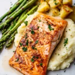Salmon dinner aesthetic - the image shows a plate of food consisting of a piece of salmon, mashed potatoes, and asparagus. the salmon is cooked to a golden brown color and is garnished with fresh herbs. the mashed potatoes are white and fluffy, and the aspartagus is a bright green color. the plate is white and the food is arranged neatly on a marble surface. the overall presentation of the dish is elegant and appetizing.