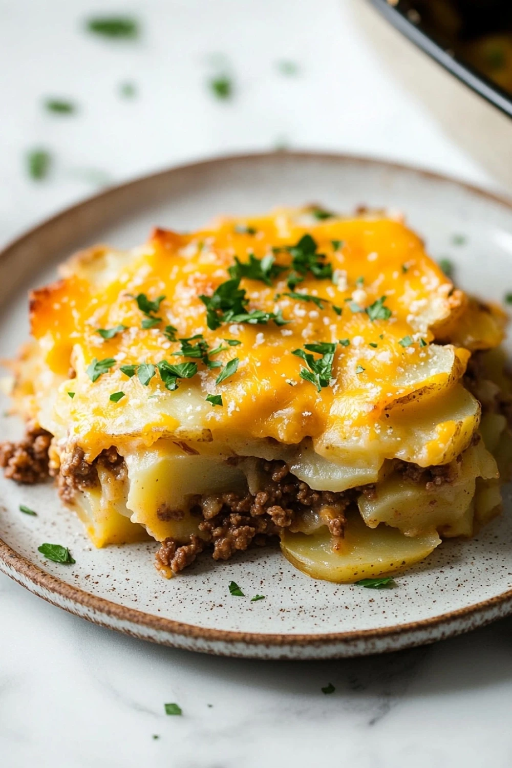 Scalloped potato casserole with ground beef - the image shows a plate of lasagna on a white marble countertop. the lasagna is layered with layers of potatoes, meat, and cheese. the top layer is covered in melted cheese and garnished with chopped parsley. the plate is round and has a light brown rim. the background is blurred, but it appears to be a kitchen countertop with a baking dish visible in the top right corner.