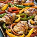 Sheet pan chicken fajitas with thighs - the image shows a baking tray filled with grilled chicken and colorful bell peppers. the chicken is golden brown and appears to be seasoned with herbs and spices. the bell peppers are of different colors - red, yellow, green, and orange - and are arranged in a circular pattern on the tray. the tray is lined with parchment paper and is sitting on a dark wooden table. the background is blurred, but it seems to be a kitchen countertop.