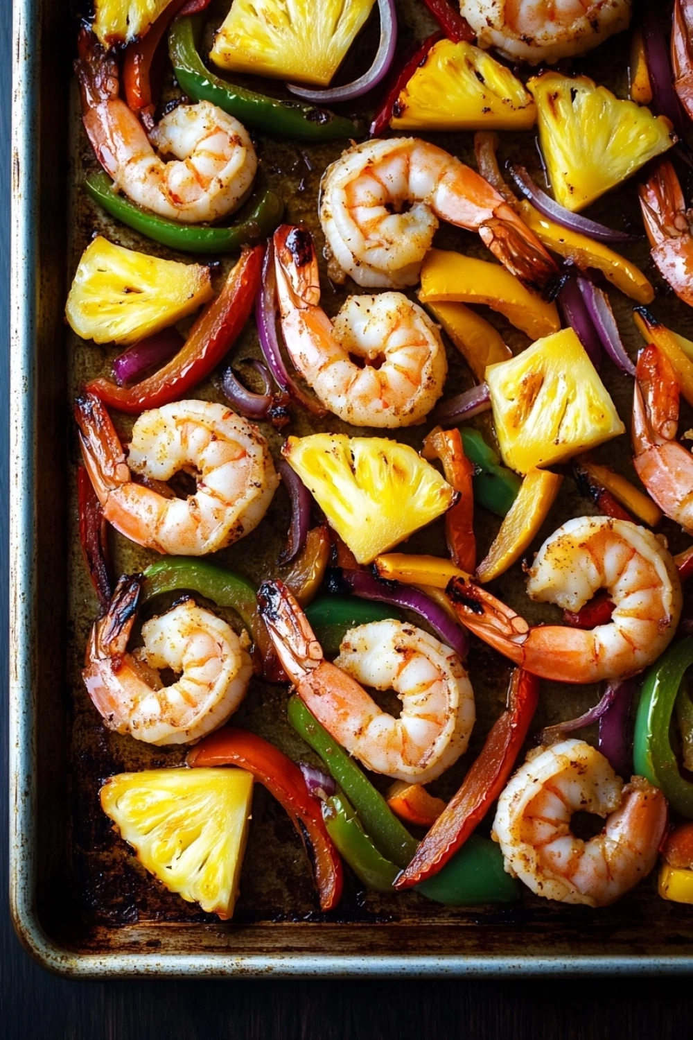 Sheet pan shrimp fajitas with pineapple - the image shows a baking tray filled with shrimp and vegetables. the shrimp are cooked medium-sized and have a pinkish-orange color. they are arranged in a circular pattern on the tray, with some overlapping each other. the vegetables include red, yellow, green, and orange bell peppers, as well as sliced pineapples and red onions. the tray is lined with parchment paper, and the background is dark. the overall appearance of the dish is colorful and appetizing.