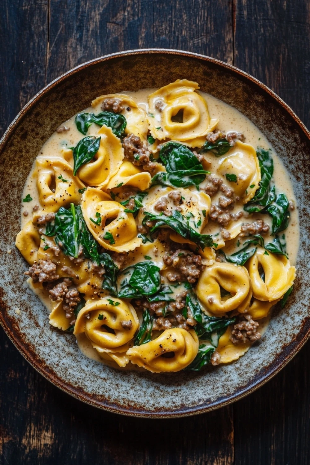 Skillet beef tortellini in creamy sauce - the image is a close-up of a dish in a brown ceramic bowl. the dish appears to be a tortellini pasta dish with a creamy sauce and chunks of ground beef and spinach mixed in. the sauce is a light beige color and is drizzled over the top of the pasta. the spinach is a bright green color and looks fresh and vibrant. the bowl is sitting on a dark wooden table.