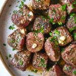 Skillet garlic butter steak bites - the image is a close-up of a dish in a bowl. the dish appears to be a type of steak or beef tenderloin, with a dark brown color and a crispy texture. the steak is garnished with chopped herbs and chopped nuts, giving it a savory flavor. the bowl is white with a light brown rim, and the background is blurred, making the dish the focal point of the image.