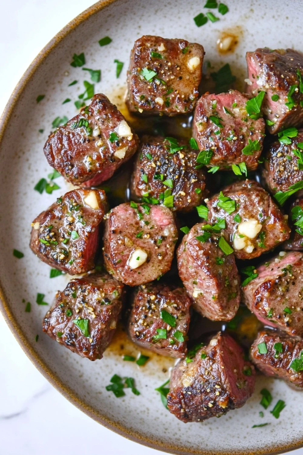 Skillet garlic butter steak bites - the image is a close-up of a dish in a bowl. the dish appears to be a type of steak or beef tenderloin, with a dark brown color and a crispy texture. the steak is garnished with chopped herbs and chopped nuts, giving it a savory flavor. the bowl is white with a light brown rim, and the background is blurred, making the dish the focal point of the image.