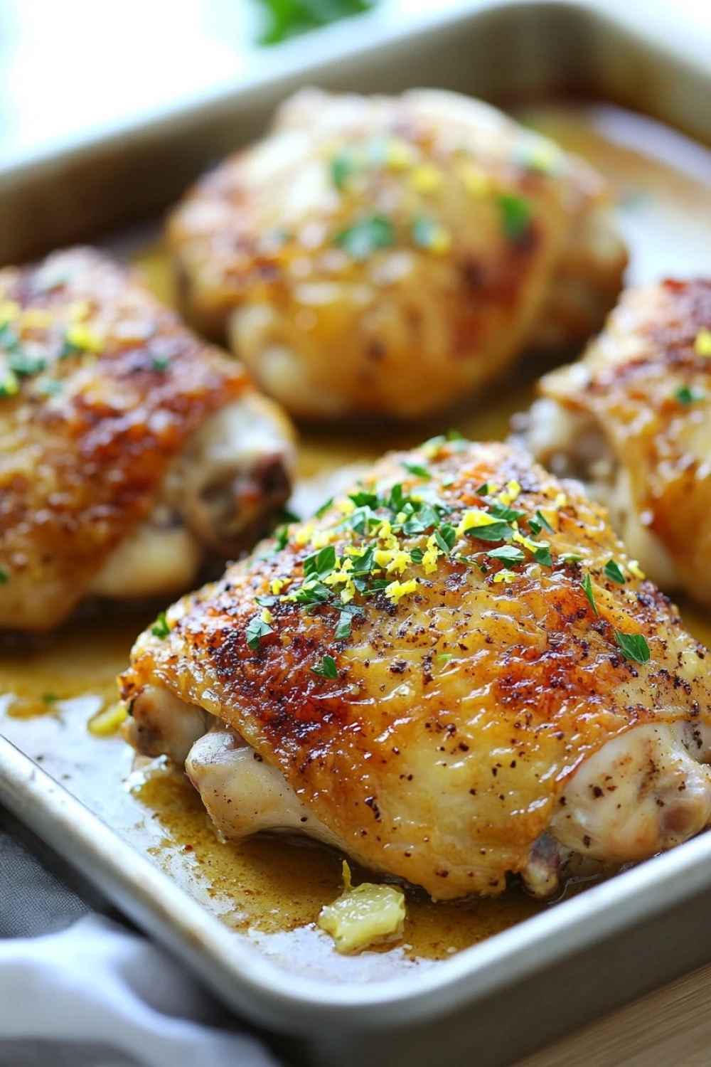 Skillet roasted chicken in lemon sauce - the image shows a baking tray with four pieces of chicken breasts. the chicken breasts are golden brown and appear to be seasoned with herbs and spices. the tray is lined with parchment paper and there is a sprig of parsley on the side. the background is blurred, but it appears to be a kitchen countertop.