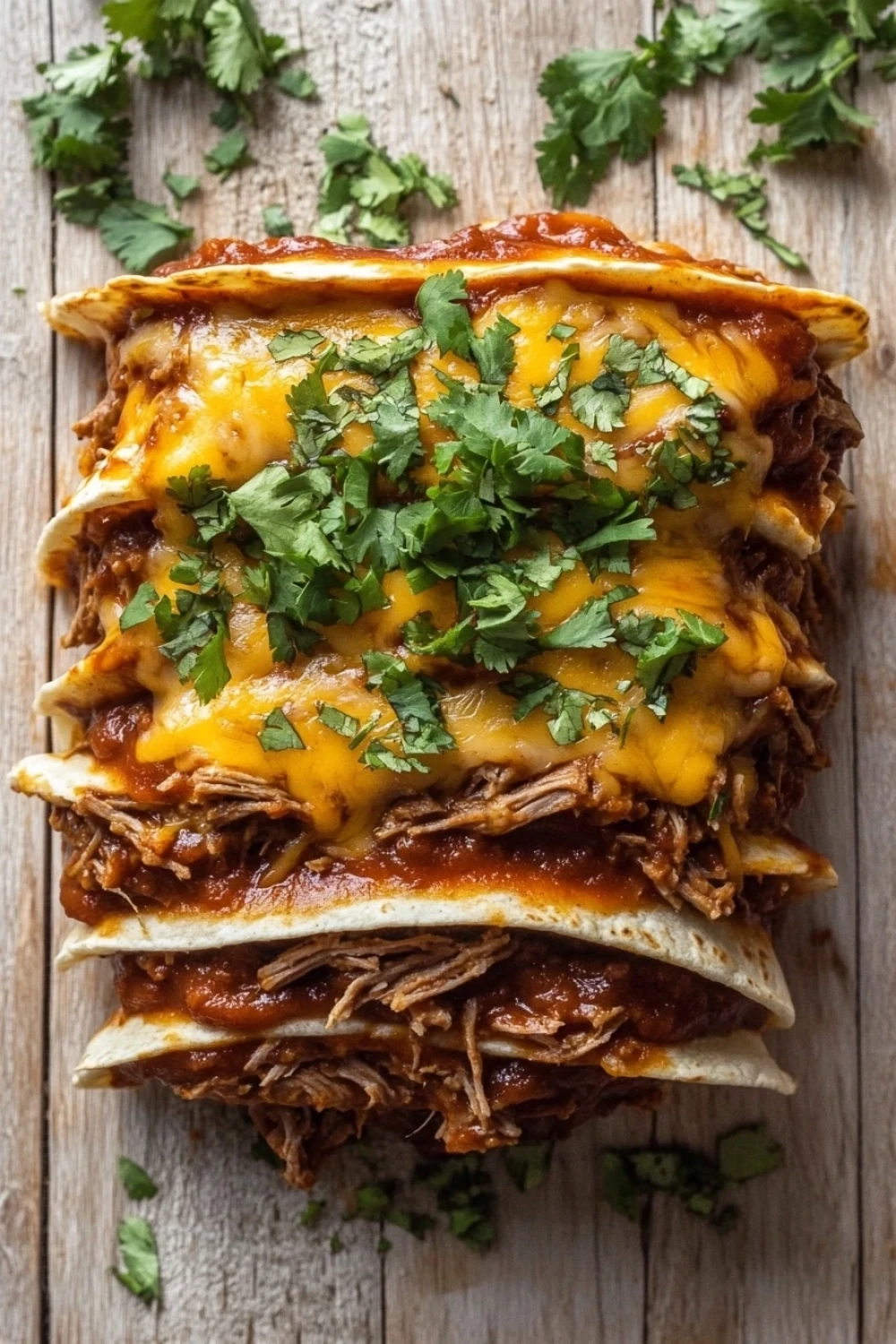 Slow cooker beef enchilada stack - the image shows a stack of three enchiladas on a wooden surface. the enchillas are golden brown and appear to be made with shredded beef and cheese. the cheese is melted and bubbly on top, and there are a few sprigs of fresh cilantro scattered around the stack. the background is a rustic wooden table.