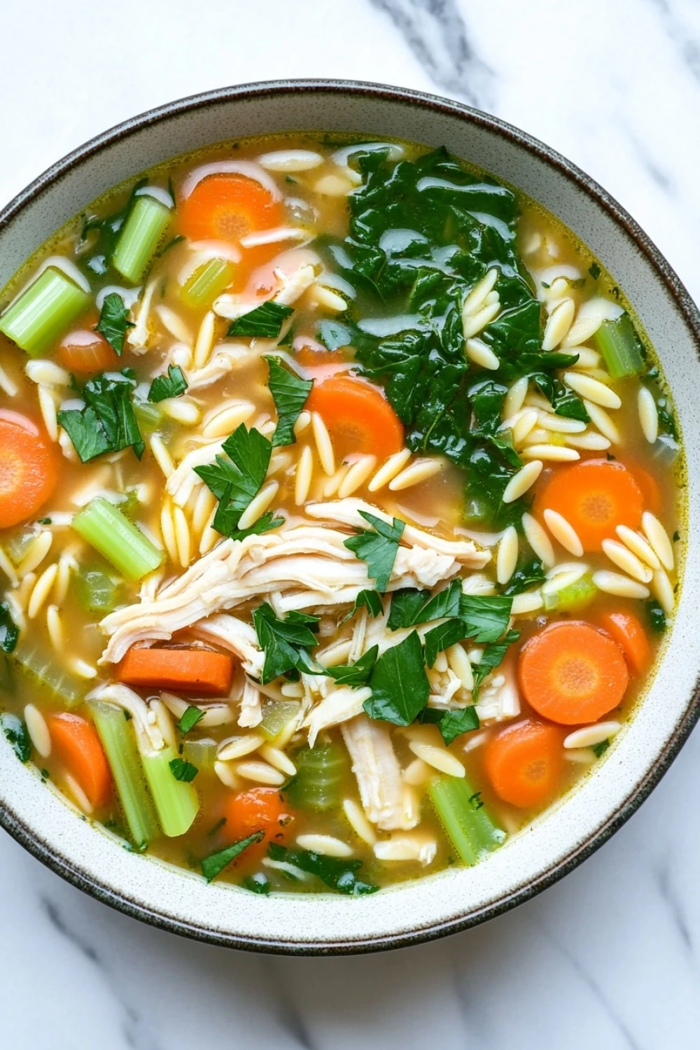Slow cooker chicken orzo soup crockpot - the image is a close-up of a bowl of soup. the soup appears to be a vegetable soup with chunks of chicken, carrots, celery, and greens mixed in. the bowl is made of ceramic and is sitting on a white marble countertop. the broth is a light yellow color and looks thick and creamy. there are also some green herbs scattered throughout the soup, adding a pop of color to the dish.