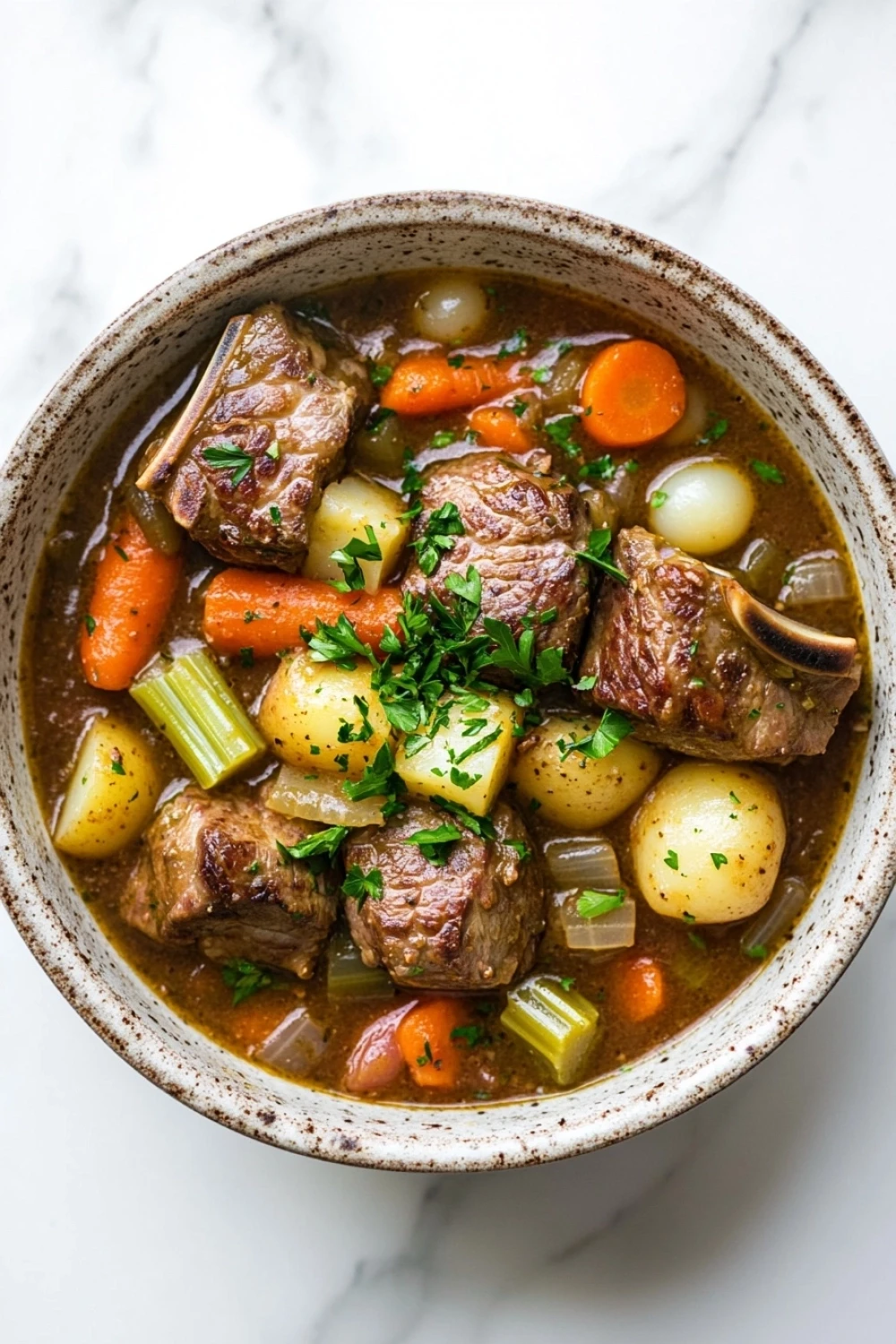Slow cooker lamb chop stew - the image is a close-up of a bowl of beef stew. the stew is made with chunks of beef, carrots, potatoes, celery, and onions. the beef is cooked medium-rare and appears to be seasoned with herbs and spices. the vegetables are bright orange and look fresh and vibrant. the bowl is made of ceramic and is sitting on a white marble countertop.