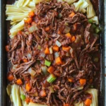 Slow cooker pot roast ragu - the image shows a rectangular black baking dish filled with a dish of beef stew. the beef appears to be cooked medium-rare and is covered in a thick, dark sauce. the dish is garnished with diced carrots, celery, and onions. the noodles are long and thin, and they are arranged in a pile on top of the beef. the sauce is a rich, dark brown color and looks thick and thick. the overall appearance of the dish is hearty and appetizing.
