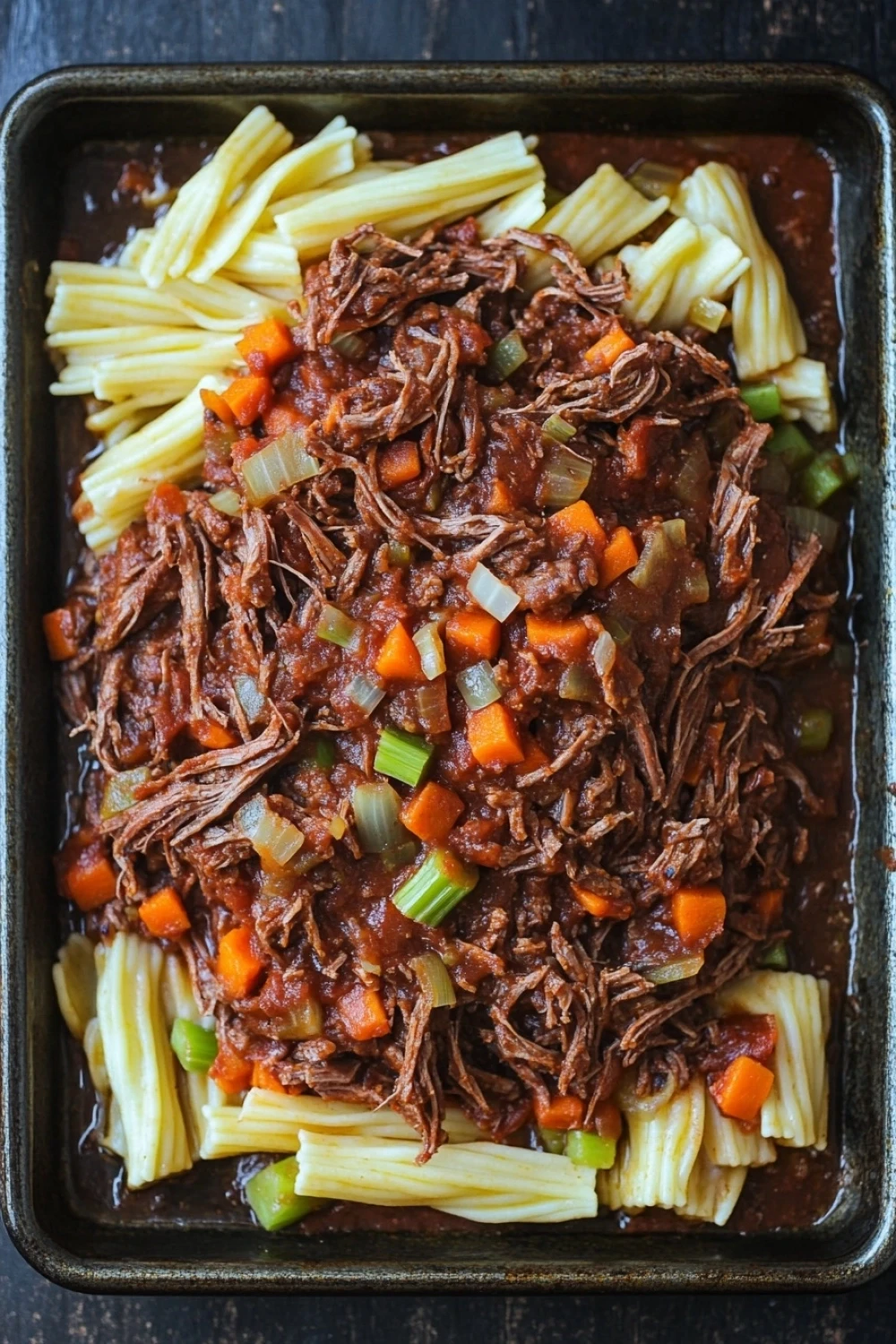 Slow cooker pot roast ragu - the image shows a rectangular black baking dish filled with a dish of beef stew. the beef appears to be cooked medium-rare and is covered in a thick, dark sauce. the dish is garnished with diced carrots, celery, and onions. the noodles are long and thin, and they are arranged in a pile on top of the beef. the sauce is a rich, dark brown color and looks thick and thick. the overall appearance of the dish is hearty and appetizing.