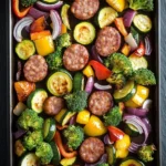 Smoked sausage veggie sheet pan - the image shows a rectangular baking tray filled with a variety of vegetables. the vegetables include zucchini, broccoli, red onions, and yellow bell peppers. there are also several sausage balls scattered throughout the tray. the tray is black and appears to be made of metal. the background is a dark grey countertop.