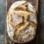Sourdough sandwich bread no yeast - the image shows a loaf of bread on a wooden cutting board. the bread appears to be freshly baked, with a golden brown crust and a soft, fluffy interior. the loaf is placed on a dark grey textured surface, possibly a countertop or a table. the edges of the bread are slightly frayed, indicating that it has been freshly baked. the overall appearance is rustic and cozy.