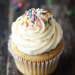 Sugar cookie cupcakes - the image is of a cupcake with white frosting on top. the cupcake is in a white paper liner and is placed on a wooden surface. the frosting is swirled in a swirl pattern and is decorated with colorful sprinkles in shades of pink, blue, green, and yellow. the sprinkles are scattered around the cupcake, adding a pop of color to the image. the background is blurred, but it appears to be a rustic wooden table.