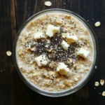 Sugar cookie overnight oats - the image is a close-up of a bowl of oatmeal on a wooden table. the oatmeal is a light beige color and appears to be creamy and smooth. it is topped with small pieces of white chocolate and sprinkled with black sesame seeds. there are also a few scattered walnuts on the table around the bowl. the bowl is made of clear glass and is sitting on a dark wooden surface.