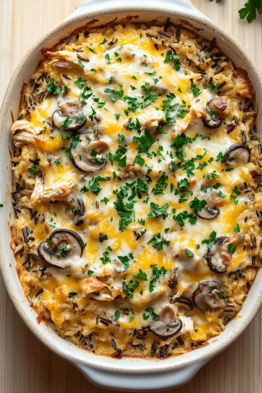 Trisha yearwood chicken wild rice casserole - the image shows a large white ceramic baking dish filled with a casserole dish. the dish appears to be made with rice, mushrooms, and chicken, and is topped with melted cheese and fresh parsley. the rice is golden brown and fluffy, and the mushrooms are scattered throughout the dish. there are also a few sprigs of parsley scattered around the dish, adding a pop of green color to the dish and giving it a fresh and appetizing appearance. the background is a wooden cutting board, making the dish stand out.
