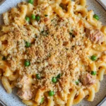 Tuna casserole cheesy - the image is a close-up of a plate of macaroni and cheese. the plate is round and has a speckled pattern around the edges. the pasta is cooked al dente and is covered in a creamy yellow sauce with chunks of chicken and peas scattered throughout. the dish is garnished with a sprinkle of breadcrumbs. the background is a light beige color, and the plate is sitting on a wooden table.