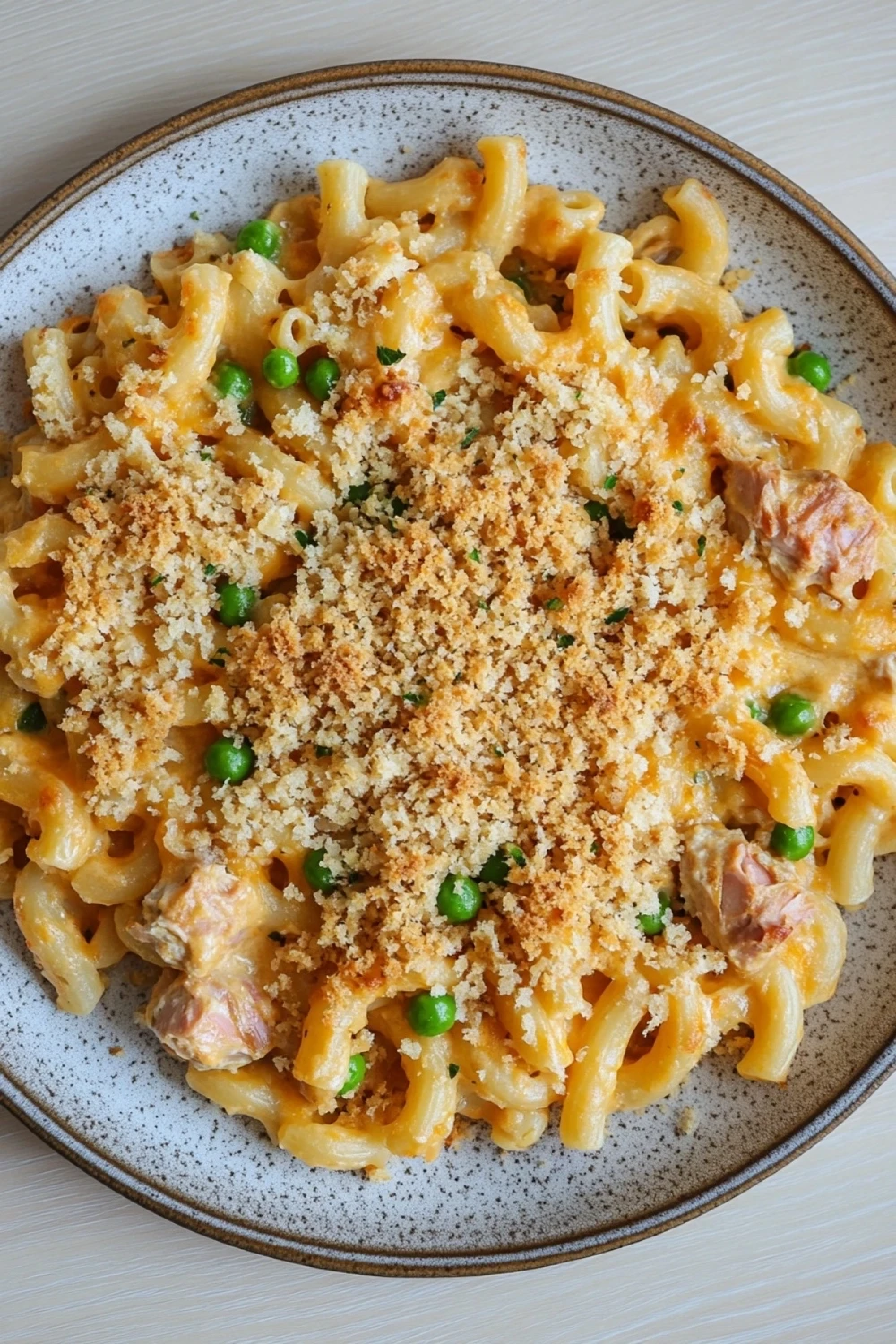 Tuna casserole cheesy - the image is a close-up of a plate of macaroni and cheese. the plate is round and has a speckled pattern around the edges. the pasta is cooked al dente and is covered in a creamy yellow sauce with chunks of chicken and peas scattered throughout. the dish is garnished with a sprinkle of breadcrumbs. the background is a light beige color, and the plate is sitting on a wooden table.