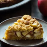 With yellow cake mix and apple pie filling - the image shows a slice of apple pie on a gray plate. the pie has a golden brown crust on top and is filled with sliced apples. the apples are arranged in a crumbly pattern and appear to be freshly baked. in the background, there is another pie and a red apple on a wooden table. the overall mood of the image is rustic and appetizing.