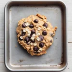 Yogurt peanut butter cookie dough - the image shows a round cookie in a stainless steel baking tray. the cookie appears to be freshly baked and has a golden brown color. it is covered in chocolate chips and oats, giving it a crunchy texture. the tray is placed on a white wooden surface.