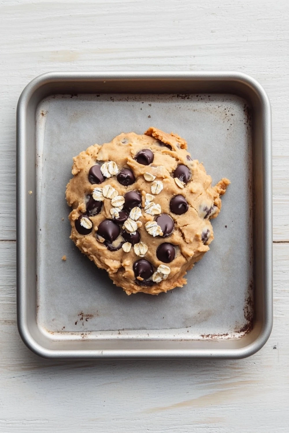 Yogurt peanut butter cookie dough - the image shows a round cookie in a stainless steel baking tray. the cookie appears to be freshly baked and has a golden brown color. it is covered in chocolate chips and oats, giving it a crunchy texture. the tray is placed on a white wooden surface.