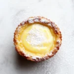 Zesty lemon bar cookie cups - the image is a close-up of a small tart on a white marble surface. the tart has a golden brown crust and is dusted with powdered sugar. in the center of the tart, there is a creamy yellow filling that appears to be a lemon curd. the filling is smooth and shiny, and the edges are slightly curled. the background is blurred, making the tart the focal point of the image.