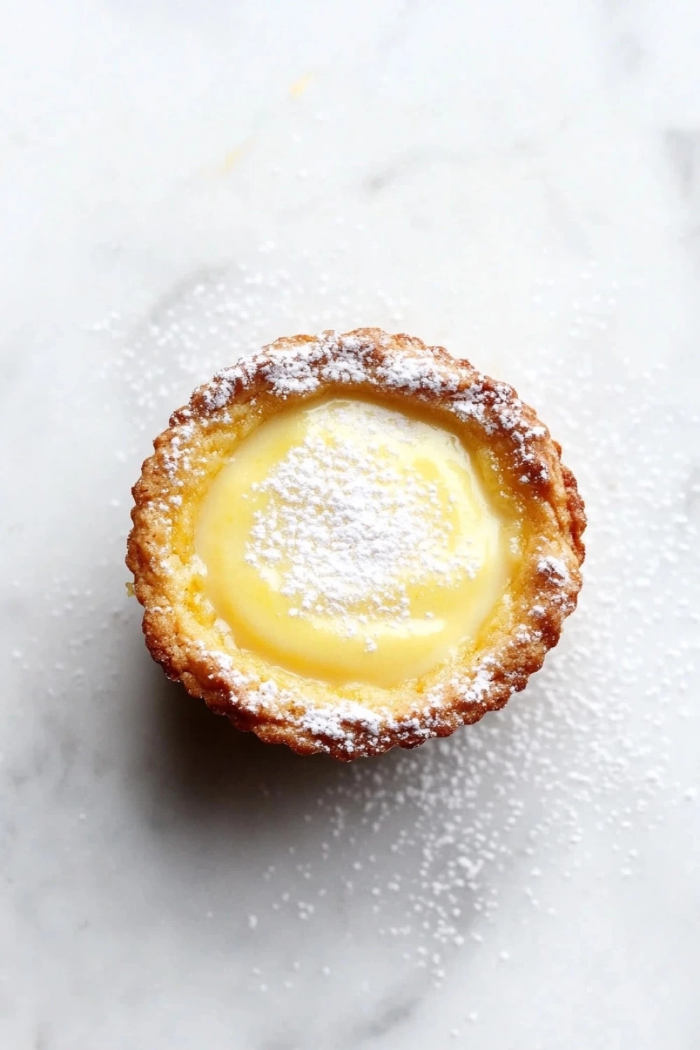 Zesty lemon bar cookie cups - the image is a close-up of a small tart on a white marble surface. the tart has a golden brown crust and is dusted with powdered sugar. in the center of the tart, there is a creamy yellow filling that appears to be a lemon curd. the filling is smooth and shiny, and the edges are slightly curled. the background is blurred, making the tart the focal point of the image.