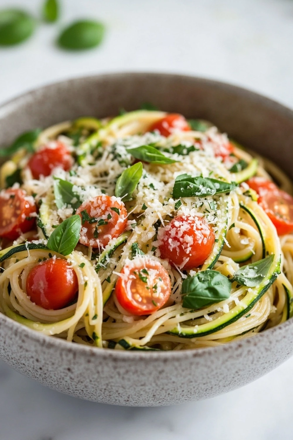 Zuchinis dinner pasta - the image is a close-up of a bowl of pasta dish. the bowl is made of ceramic and is placed on a white marble countertop. the pasta appears to be fettuccine alfredo, which is a type of italian pasta. it is topped with sliced zucchini, cherry tomatoes, and fresh basil leaves. the tomatoes are bright red and appear to be ripe and juicy. the basil leaves are scattered throughout the dish, adding a pop of green color. the dish is garnished with grated parmesan cheese, giving it a fresh and appetizing appearance.