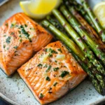 Air fryer salmon and asparagus - the image shows two pieces of salmon on a plate with asparagus. the salmon is cooked to a golden brown color and is garnished with herbs and lemon wedges. the plate is white with a speckled pattern and is placed on a dark grey countertop. the asparaglips are arranged on the side of the plate, with some of them overlapping each other. the overall presentation of the dish is elegant and appetizing.