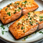 Air fryer salmon garlic - the image shows two pieces of salmon on a white plate. the salmon is cooked to a golden brown color and appears to be seasoned with herbs and spices. the plate is garnished with chopped parsley and pine nuts. the background is blurred, but it seems to be a kitchen countertop.