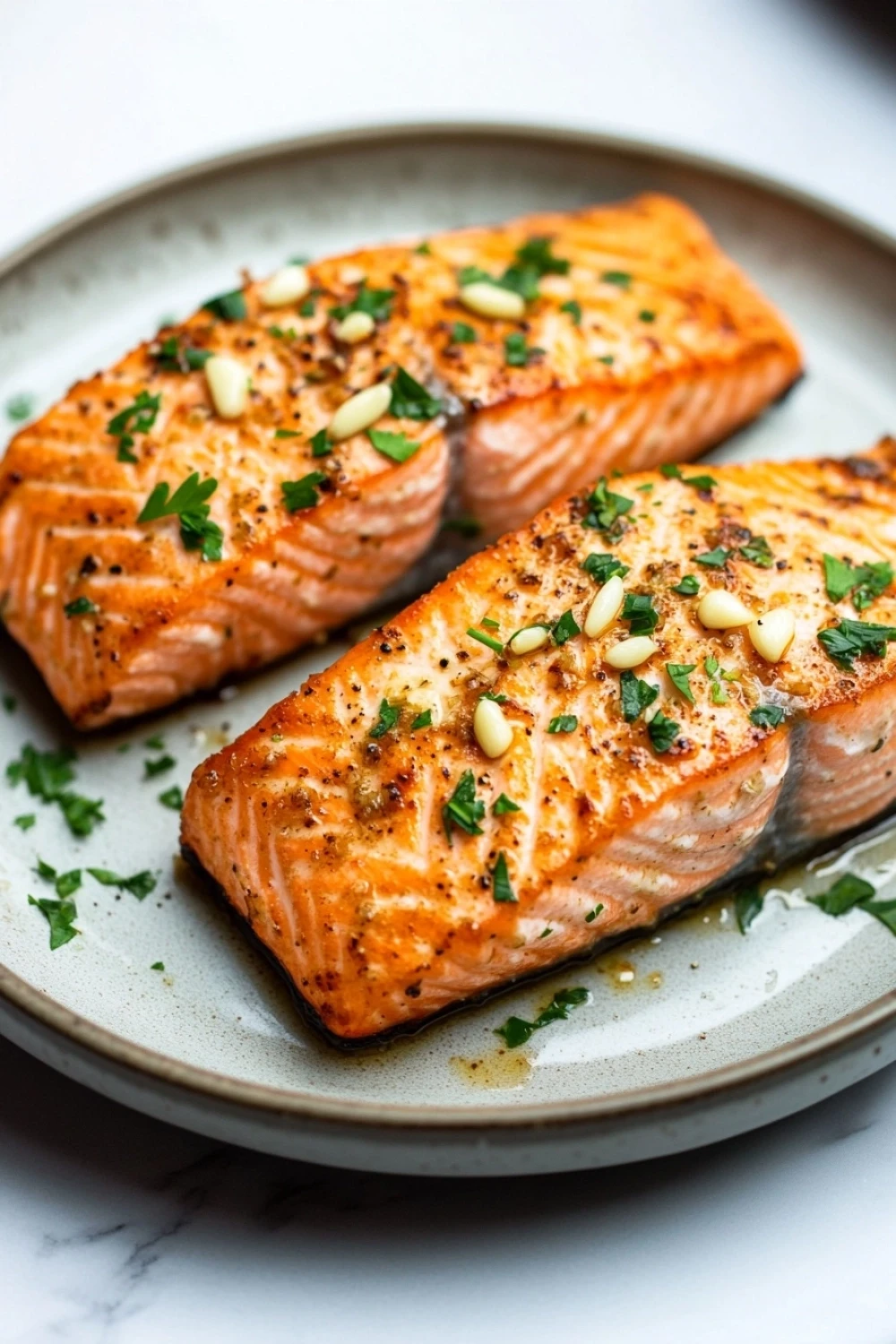 Air fryer salmon garlic - the image shows two pieces of salmon on a white plate. the salmon is cooked to a golden brown color and appears to be seasoned with herbs and spices. the plate is garnished with chopped parsley and pine nuts. the background is blurred, but it seems to be a kitchen countertop.