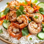 Air fryer shrimp rice bowl - the image is a close-up of a plate of food. the plate is made of a light-colored ceramic bowl with a speckled pattern. on top of the rice, there are several large shrimp arranged in a circular pattern. the shrimp are cooked to a golden brown color and appear to be seasoned with herbs and spices. the rice is white and fluffy, and there are sliced cucumbers, carrots, and red bell peppers scattered throughout the dish. the dish is garnished with fresh cilantro leaves. the background is a white marble countertop.