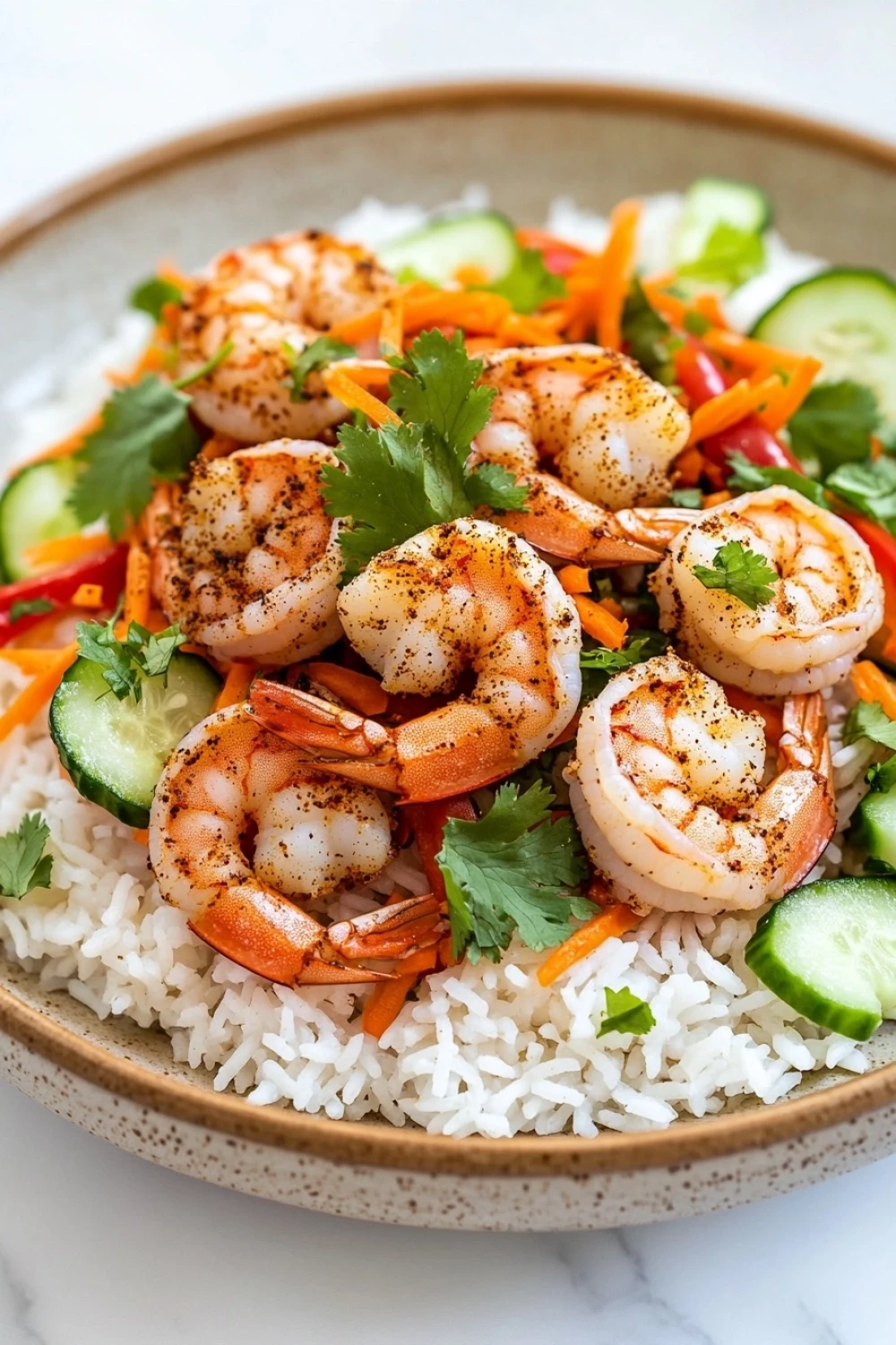Air fryer shrimp rice bowl - the image is a close-up of a plate of food. the plate is made of a light-colored ceramic bowl with a speckled pattern. on top of the rice, there are several large shrimp arranged in a circular pattern. the shrimp are cooked to a golden brown color and appear to be seasoned with herbs and spices. the rice is white and fluffy, and there are sliced cucumbers, carrots, and red bell peppers scattered throughout the dish. the dish is garnished with fresh cilantro leaves. the background is a white marble countertop.
