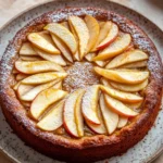 Apple cake german - the image is of a round cake on a white plate. the cake is golden brown in color and has a crumbly texture. on top of the cake, there are sliced apples arranged in a circular pattern. the apples are cut into thin slices and are dusted with powdered sugar. the plate is sitting on a beige countertop with a few other plates in the background.