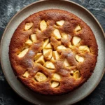 Apple cake kids - the image is a close-up of a round cake on a gray plate. the cake appears to be freshly baked and has a golden brown crust. on top of the cake, there are small pieces of sliced apples arranged in a criss-cross pattern. the apples are cut into small cubes and are scattered across the cake. the plate is placed on a dark, textured surface. the overall mood of the image is rustic and appetizing.