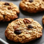 Baked oatmeal cookie and kate - the image shows a tray of freshly baked oatmeal cookies. the cookies are golden brown and have a crumbly texture. they are topped with chopped pecans and raisins, giving them a crunchy appearance. the tray is lined with parchment paper, and the background is dark, making the cookies stand out.