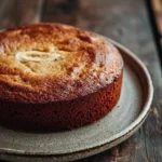 Banana cake from scratch - the image is a close-up of a round cake on a gray plate. the cake appears to be freshly baked and has a golden brown crust on top. the top of the cake is covered in a light dusting of powdered sugar, giving it a shiny appearance. the plate is placed on a wooden table with a rustic, weathered look. the background is blurred, making the cake the focal point of the image.