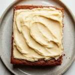 Banana cake sheet cake - the image shows a square piece of cake on a gray plate. the cake appears to be a chocolate cake with a golden brown crust. on top of the cake, there is a generous layer of creamy cream cheese frosting. the frosting is spread evenly across the cake and has a smooth texture. the plate is sitting on a white marble countertop.