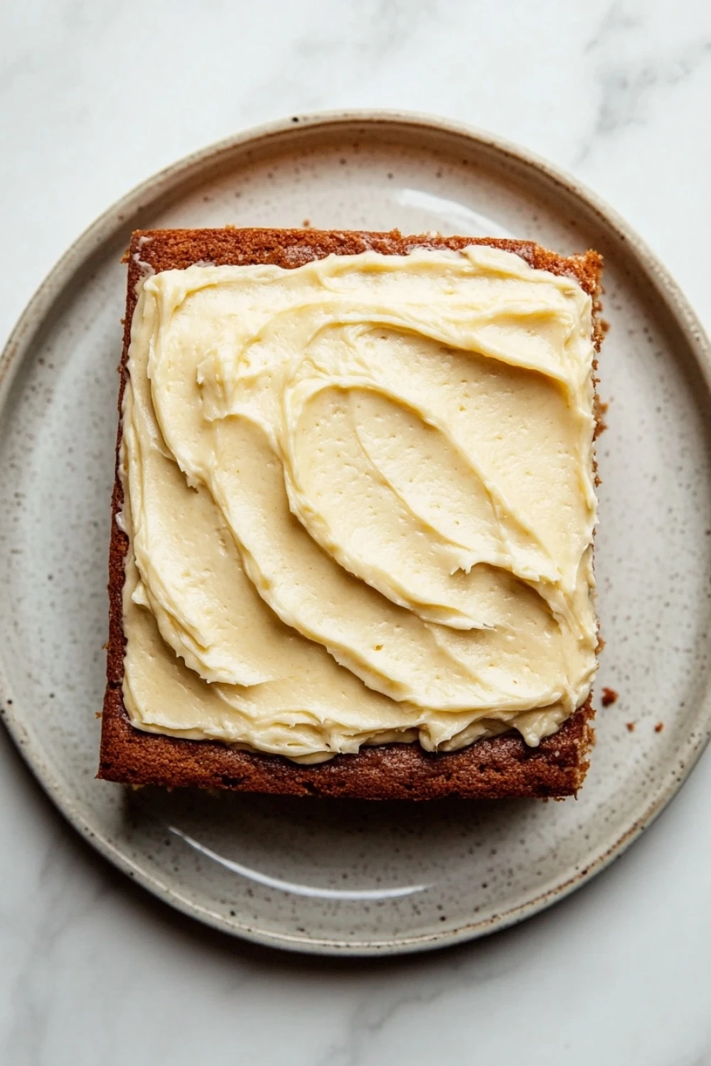 Banana cake sheet cake - the image shows a square piece of cake on a gray plate. the cake appears to be a chocolate cake with a golden brown crust. on top of the cake, there is a generous layer of creamy cream cheese frosting. the frosting is spread evenly across the cake and has a smooth texture. the plate is sitting on a white marble countertop.