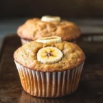 Banana muffin nut free - the image shows two freshly baked banana muffins on a wooden tray. the muffins are golden brown in color and have a crumbly texture. on top of the muffins, there are sliced bananas arranged in a circular pattern. the background is blurred, but it appears to be a rustic kitchen countertop. the overall mood of the image is warm and inviting.