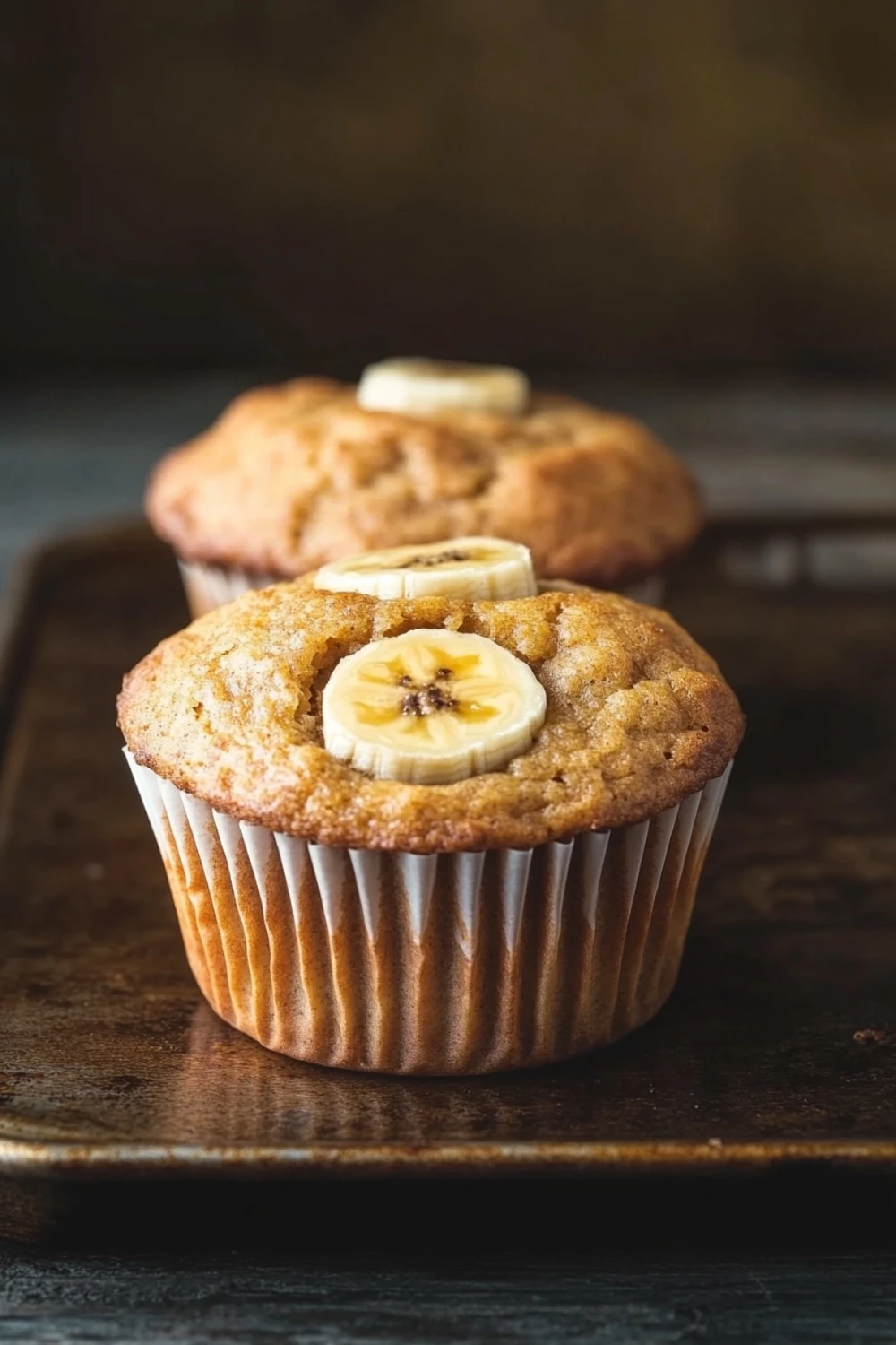 Banana muffin nut free - the image shows two freshly baked banana muffins on a wooden tray. the muffins are golden brown in color and have a crumbly texture. on top of the muffins, there are sliced bananas arranged in a circular pattern. the background is blurred, but it appears to be a rustic kitchen countertop. the overall mood of the image is warm and inviting.