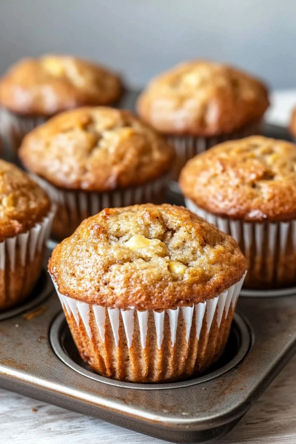 Banana muffin pictures - the image shows a group of freshly baked muffins in a muffin tin. the muffins are golden brown in color and have a crumbly texture. they are arranged in a single layer on the tray, with some overlapping each other. the tray is placed on a wooden surface, and the background is blurred, making the muffins the focal point of the image.