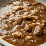 Beef chicken gravy - the image is a close-up of a bowl of soup. the soup appears to be a rich, dark brown color and has chunks of meat and vegetables mixed in. the dish is garnished with chopped nuts, giving it a crunchy texture. the bowl is made of ceramic with a speckled pattern around the edges. the background is a white marble countertop.