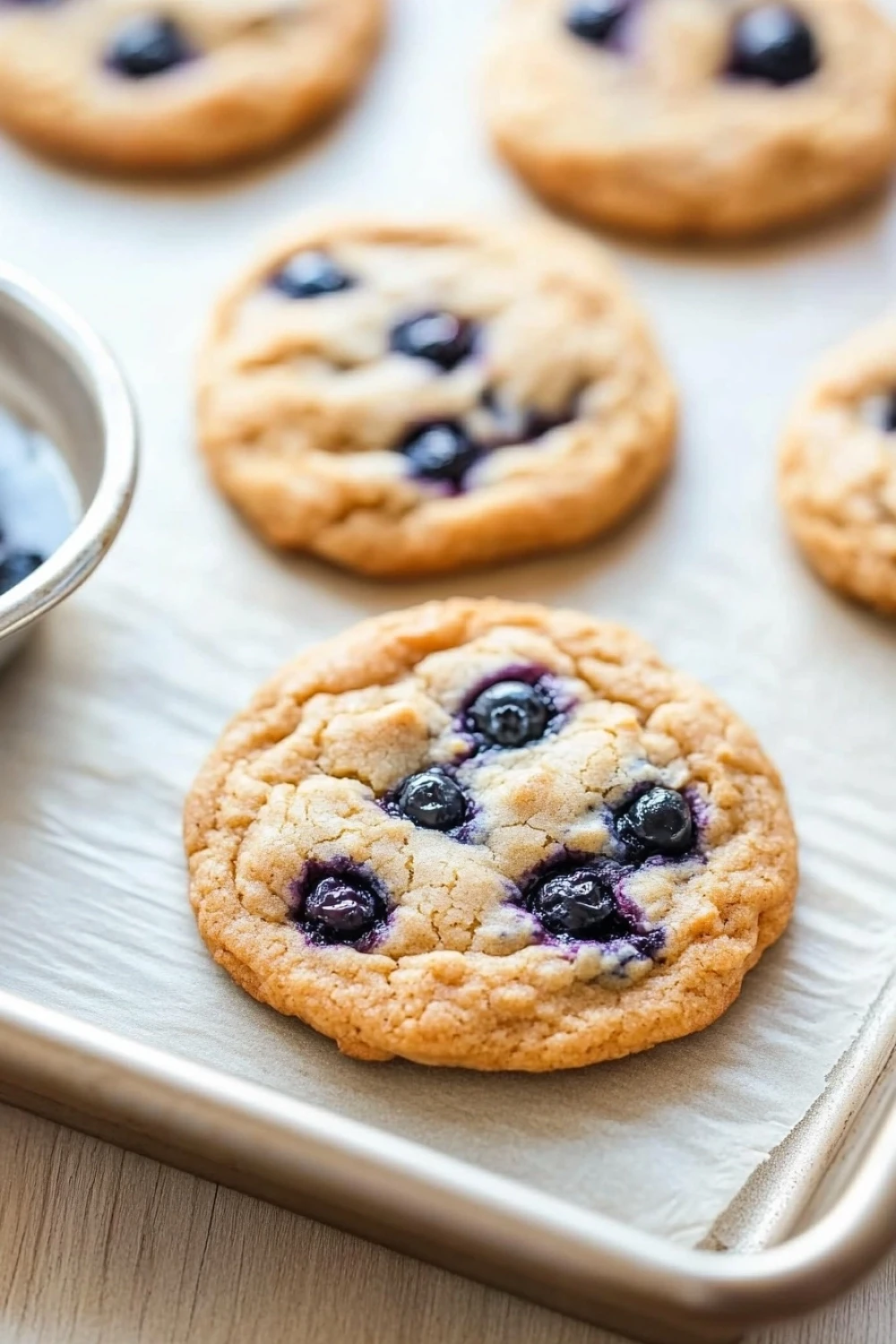 Blueberry muffin mix cookies - the image shows a baking tray with freshly baked blueberry cookies on it. the cookies are golden brown and have a crumbly texture. they are arranged in a single layer on the tray, with some of the cookies overlapping each other. there is a small bowl of blueberries on the left side of the tray. the tray is lined with parchment paper, and the background is a wooden table.