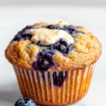 Blueberry muffin with cream cheese - the image is of a freshly baked blueberry muffin on a white marble countertop. the muffin has a golden brown crust and is topped with fresh blueberries and a dollop of white cream cheese frosting. there are two whole blueberries on the countertop next to the muffin. the background is blurred, making the blueberries the focal point of the image.